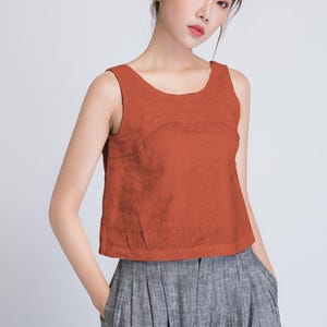 Könnte beinhalten: Ein ärmelloses, rostfarbenes Oberteil mit rundem Ausschnitt. Das Oberteil wird mit grauen, plissierten Hosen kombiniert. Das Outfit ist schlicht und elegant, geeignet für lässige oder halbformelle Anlässe. Der Stoff scheint Leinen oder ein ähnliches atmungsaktives Material zu sein.