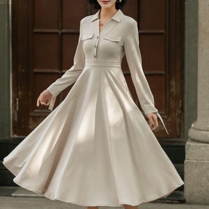 May include: A beige long-sleeved dress with a collared neckline, button closure, and a flared skirt. The dress has two pockets on the chest and a tie detail at the cuffs.