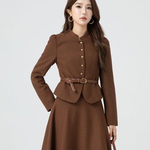 May include: A brown wool skirt suit with a fitted jacket and a flared skirt. The jacket has a mandarin collar, gold buttons, and a brown leather belt.