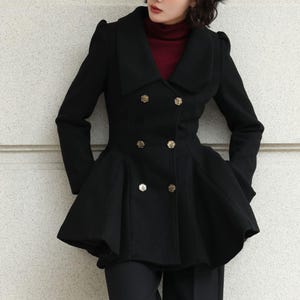 May include: A black double-breasted coat with gold buttons and a flared peplum. The coat has a wide collar and is worn over a burgundy turtleneck. The model is also wearing black trousers.