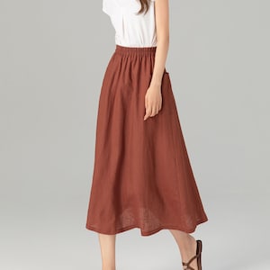 May include: A woman wearing a white t-shirt and a rust-coloured linen skirt with a side pocket. She is wearing brown sandals.