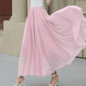 May include: A woman in a white scoop-neck t-shirt and a flowing, light pink maxi skirt. She is also wearing a white hat and white high-heeled sandals. The skirt appears to be made of a lightweight fabric.