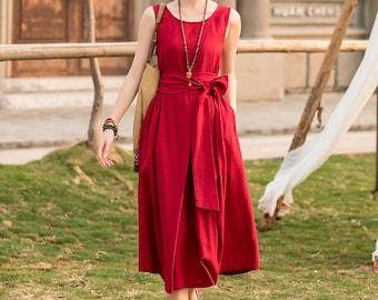 May include: A woman wearing a red sleeveless linen dress with a bow at the waist. She is walking on a grassy path with a building in the background.