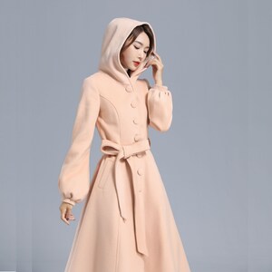 May include: A long, blush-colored coat with a hood, a button-up front, and a matching belt. The coat has a flared skirt and long sleeves with puffed cuffs.