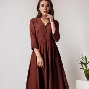 Könnte beinhalten: Ein braunes Midikleid mit V-Ausschnitt und Knopfleiste. Das Kleid hat einen plissierten Rock und 3/4 Ärmel.