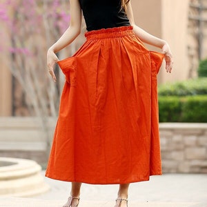 May include: A black tank top paired with a long, vibrant orange skirt. The skirt has a high waist and a flowing, pleated design. The outfit is completed with simple sandals, creating a casual yet stylish look.