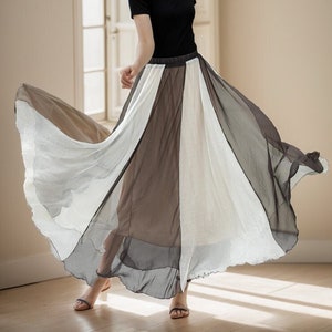 May include: A woman wearing a black top and a long, flowing skirt with alternating panels of white, grey, and brown fabric. The skirt is gathered at the waist and has a flowing, layered design.
