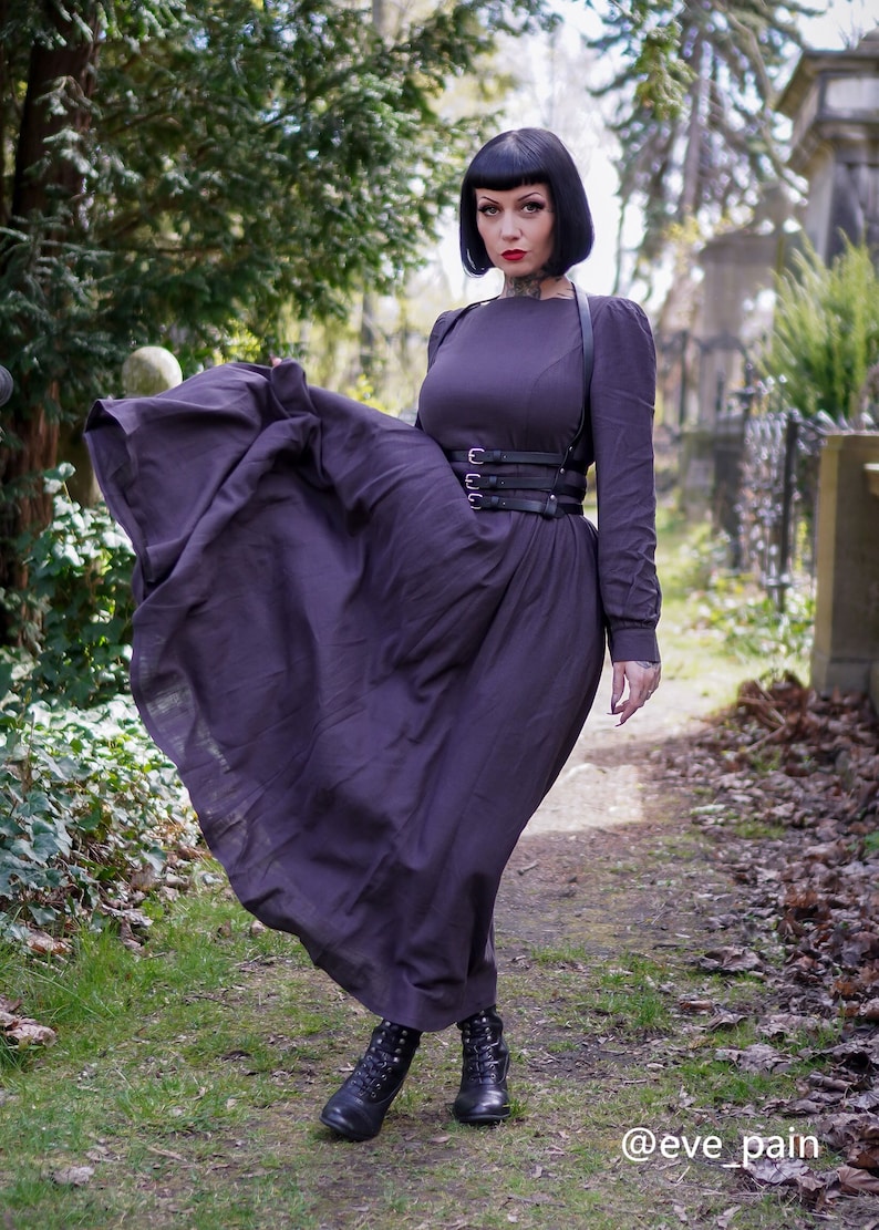 May include: A woman wearing a long, dark purple dress with a black leather harness and belt. She is standing in a cemetery with green grass and trees in the background. The text "@eve_pain" is visible in the lower right corner of the image.