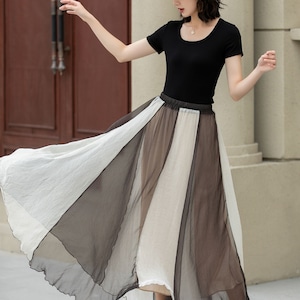 May include: A woman wearing a black top and a white hat is wearing a long, flowing skirt with alternating panels of white and brown sheer fabric.