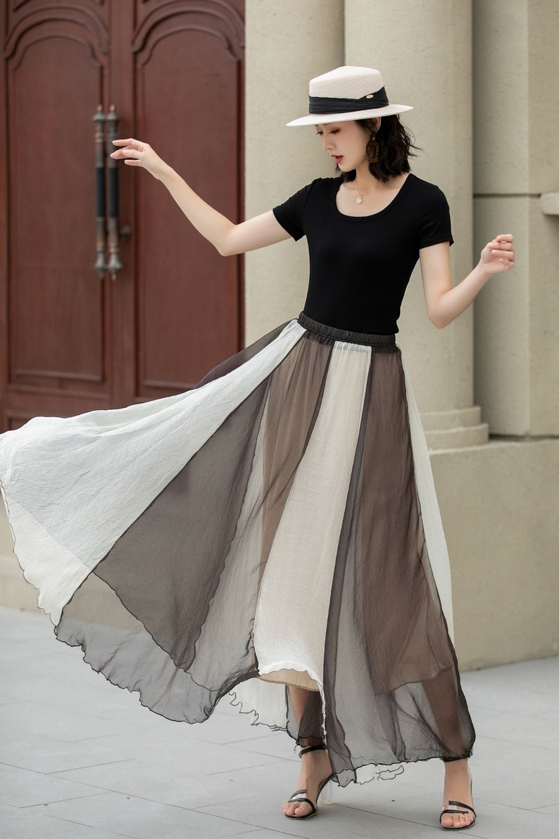 May include: A woman wearing a black top and a white hat is wearing a long, flowing skirt with alternating panels of white and brown sheer fabric.