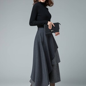 May include: A woman wearing a black turtleneck sweater and a gray layered skirt. She is holding a black clutch purse and is wearing black heels.