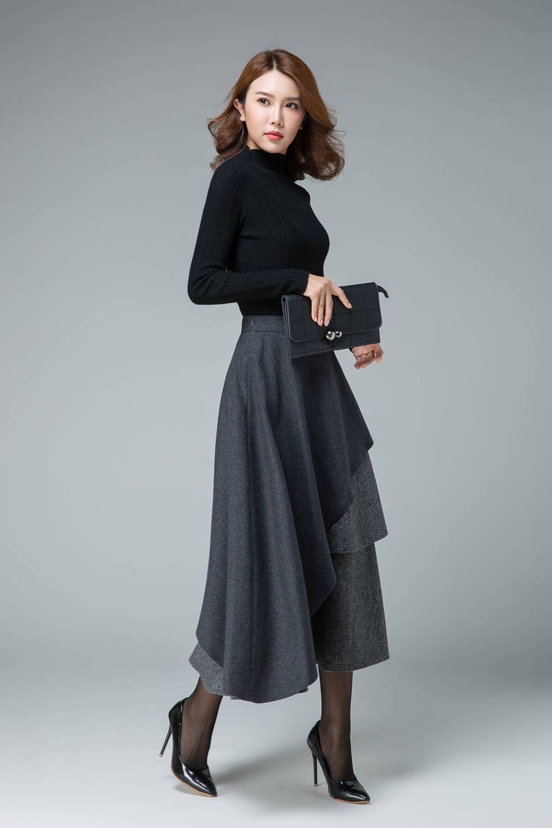 May include: A woman wearing a black turtleneck sweater and a gray layered skirt. She is holding a black clutch purse and is wearing black heels.
