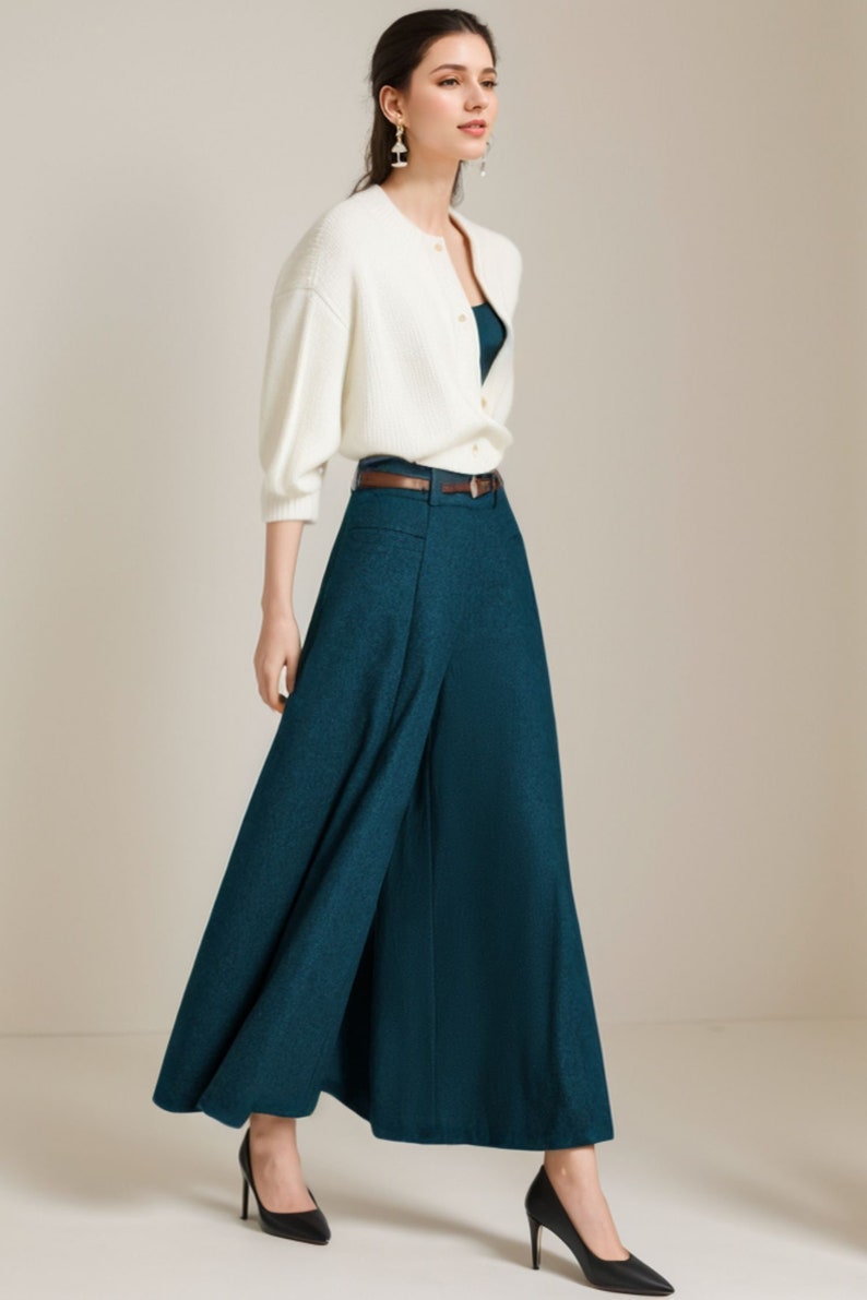 May include: A woman wearing a white cardigan, a teal wide-leg pant, and black heels. The pants have a brown belt.