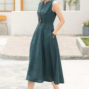 Peut inclure: Une femme portant une robe midi sans manches bleu sarcelle avec un col en V et un collier de perles. Elle est debout sur un trottoir avec un immeuble blanc en arri&egrave;re-plan.