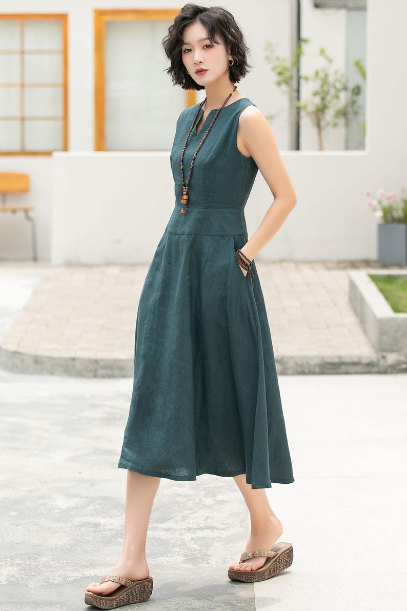 Peut inclure: Une femme portant une robe midi sans manches bleu sarcelle avec un col en V et un collier de perles. Elle est debout sur un trottoir avec un immeuble blanc en arri&egrave;re-plan.