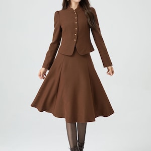 May include: A brown skirt suit with a fitted jacket and a flared skirt. The jacket has a mandarin collar and gold buttons. The skirt is knee-length and has a slight A-line shape.