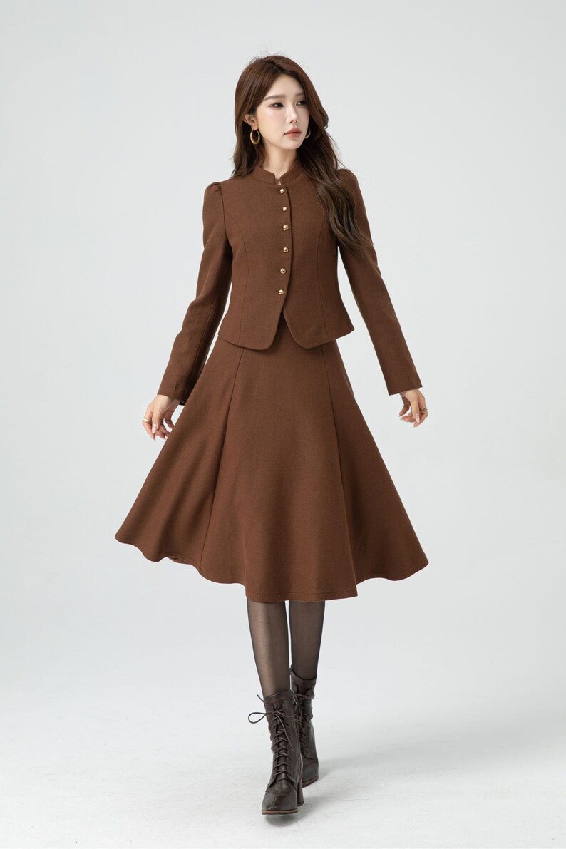 May include: A brown skirt suit with a fitted jacket and a flared skirt. The jacket has a mandarin collar and gold buttons. The skirt is knee-length and has a slight A-line shape.