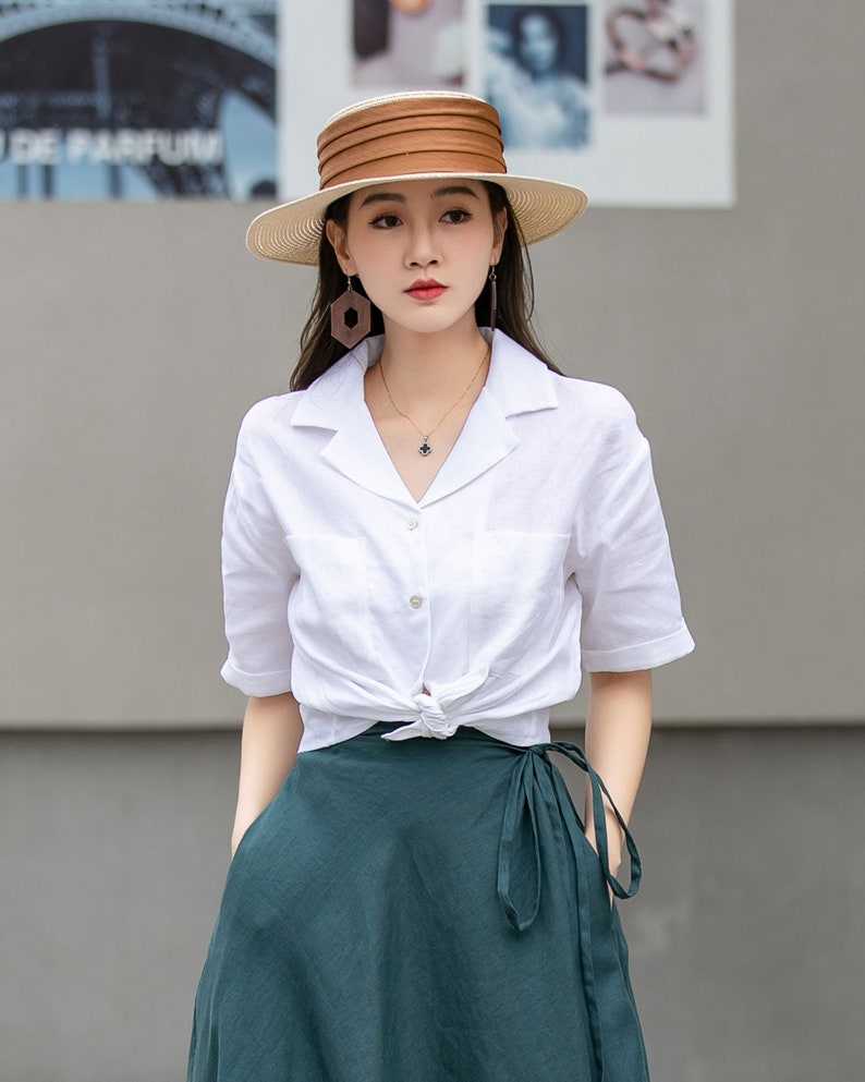 May include: A woman wearing a white linen shirt tied at the waist, a dark green wrap skirt, and a wide-brimmed straw hat with a brown band.