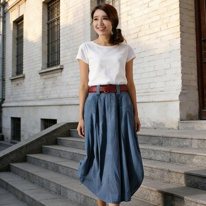 May include: A woman wearing a white short-sleeved top, a blue skirt, and a brown belt. She is standing on a set of stone steps. The skirt is a midi length and has a unique, rounded shape. She is wearing beige sandals.