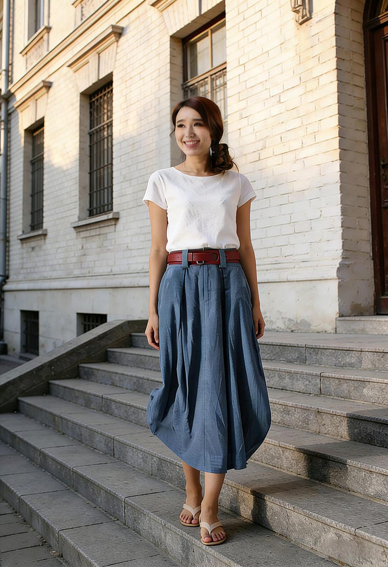 May include: A woman wearing a white short-sleeved top, a blue skirt, and a brown belt. She is standing on a set of stone steps. The skirt is a midi length and has a unique, rounded shape. She is wearing beige sandals.