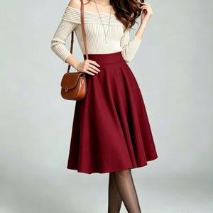 May include: A beige off-the-shoulder ribbed long-sleeve top paired with a high-waisted solid dark red midi skirt. The outfit includes a brown leather crossbody bag, sheer black tights, and black Mary Jane style shoes with a thin ankle strap.