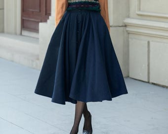 May include: A dark blue, button-down skirt with a flared silhouette. The skirt is paired with a sweater vest and a long-sleeved shirt. The image is taken outdoors.