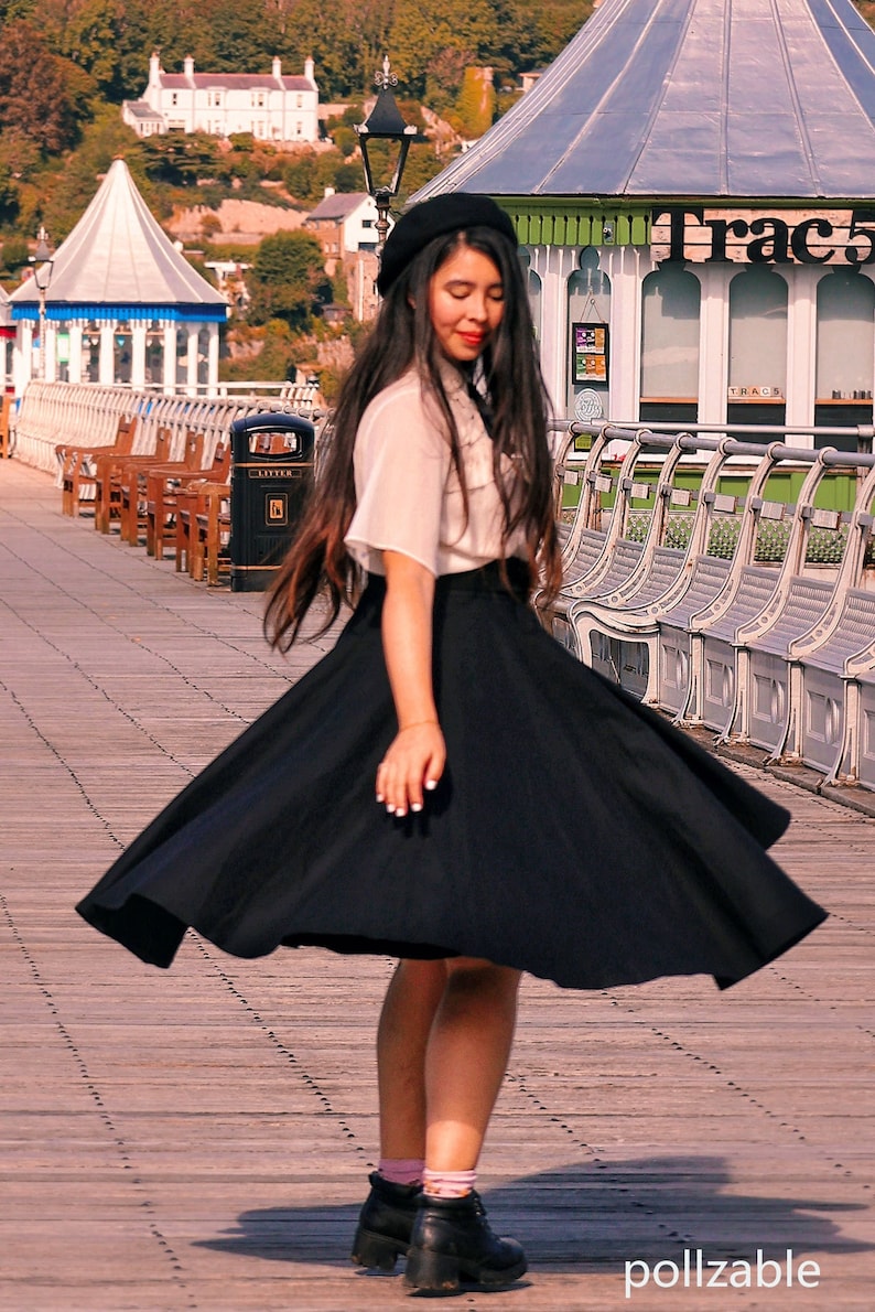 Peut inclure: Une femme portant une blouse blanche, un b&eacute;ret noir et une jupe noire se tient sur une jet&eacute;e en bois. Sa jupe flotte au vent. Elle porte des bottes noires et a de longs cheveux noirs.