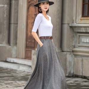 May include: A woman wearing a gray, floor-length skirt, a white top, and a brown belt. She is also wearing a black hat and black heels. The outfit is suitable for a formal or semi-formal occasion.