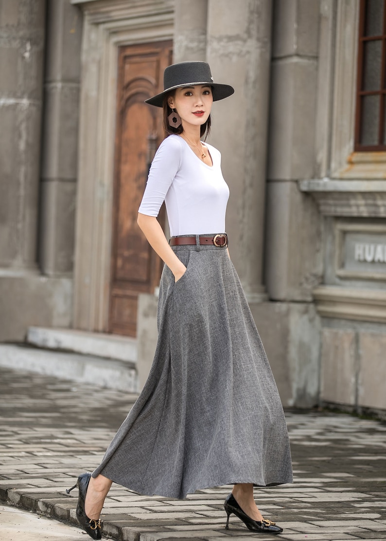 May include: A woman wearing a gray, floor-length skirt, a white top, and a brown belt. She is also wearing a black hat and black heels. The outfit is suitable for a formal or semi-formal occasion.