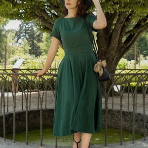 May include: A woman wearing a green linen dress with a wide, black brimmed hat and black heeled sandals. She is standing in front of a wrought iron fence and a small pond.