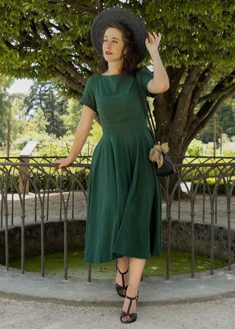 May include: A woman wearing a green linen dress with a wide, black brimmed hat and black heeled sandals. She is standing in front of a wrought iron fence and a small pond.