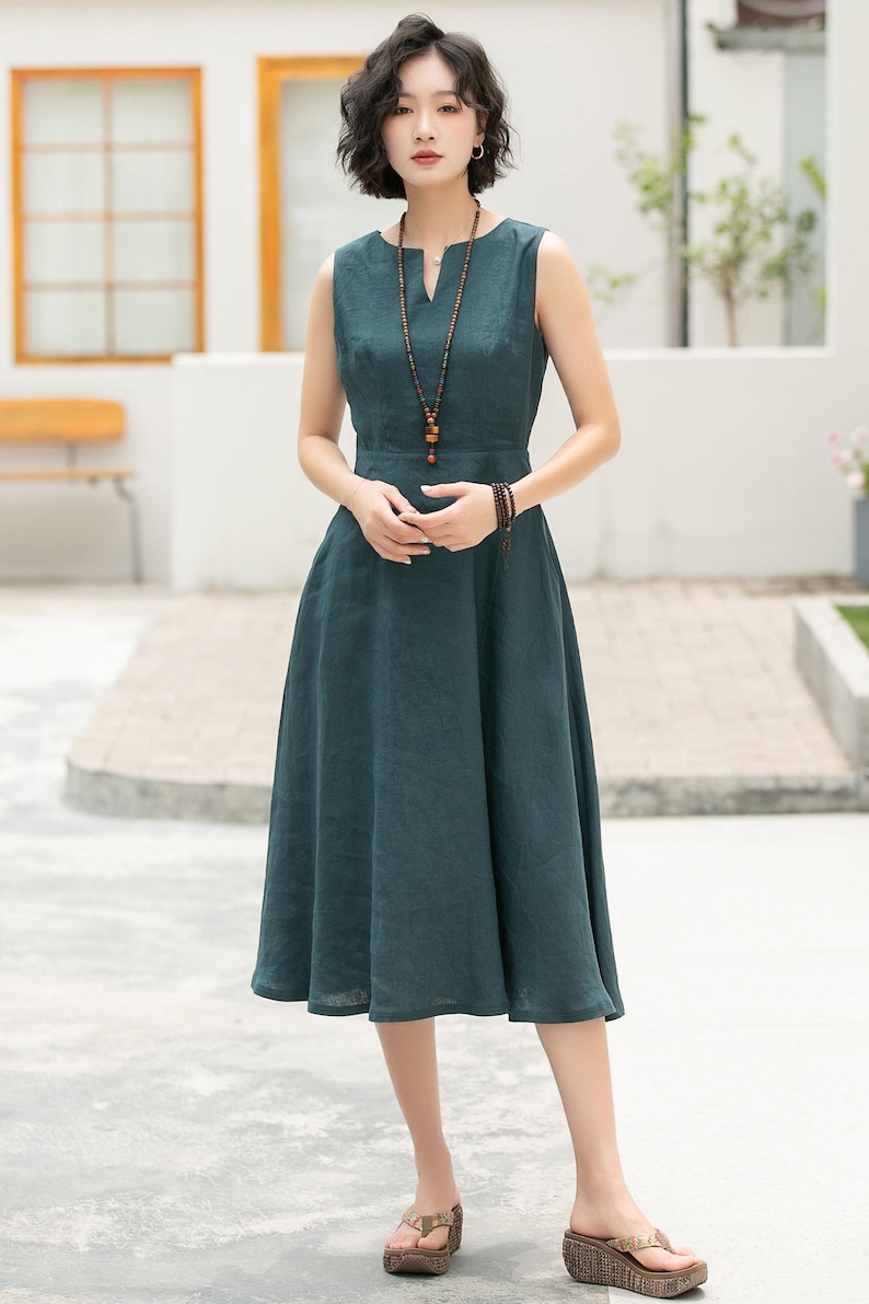 Peut inclure: Une femme portant une robe en lin sans manches bleu sarcelle avec un col en V et un collier de perles. Elle se tient sur un patio en b&eacute;ton avec un banc en bois en arri&egrave;re-plan.