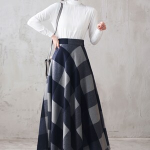 May include: A woman wearing a white long-sleeved blouse and a gray and black plaid skirt. The skirt is a-line and falls to her ankles. She is wearing black heels.