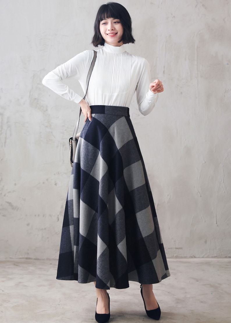 May include: A woman wearing a white long-sleeved blouse and a gray and black plaid skirt. The skirt is a-line and falls to her ankles. She is wearing black heels.