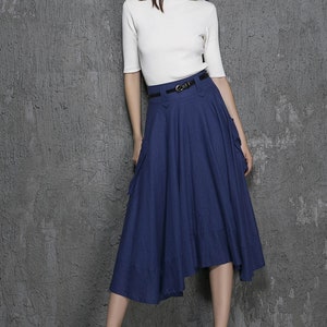 Op de afbeelding: Een vrouw draagt een wit, kort mouwig shirt en een donkerblauwe, asymmetrische rok met een zwarte riem. De rok heeft twee zakken aan de zijkanten.