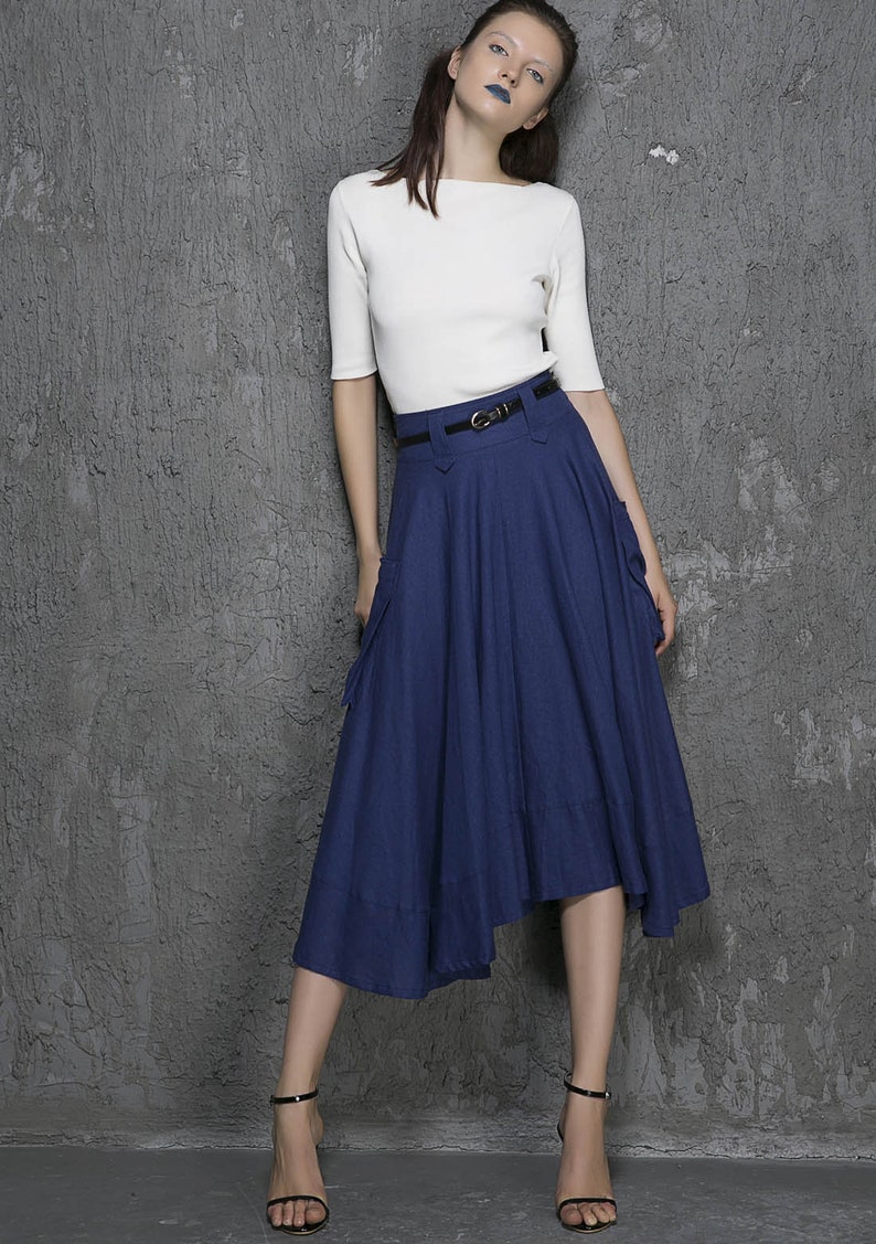 Op de afbeelding: Een vrouw draagt een wit, kort mouwig shirt en een donkerblauwe, asymmetrische rok met een zwarte riem. De rok heeft twee zakken aan de zijkanten.