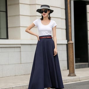 May include: A woman's outfit featuring a white scoop-neck t-shirt, a navy blue maxi skirt, and a brown belt. The outfit is accessorized with a black hat, sunglasses, and sandals. The skirt has a flowing, A-line silhouette.