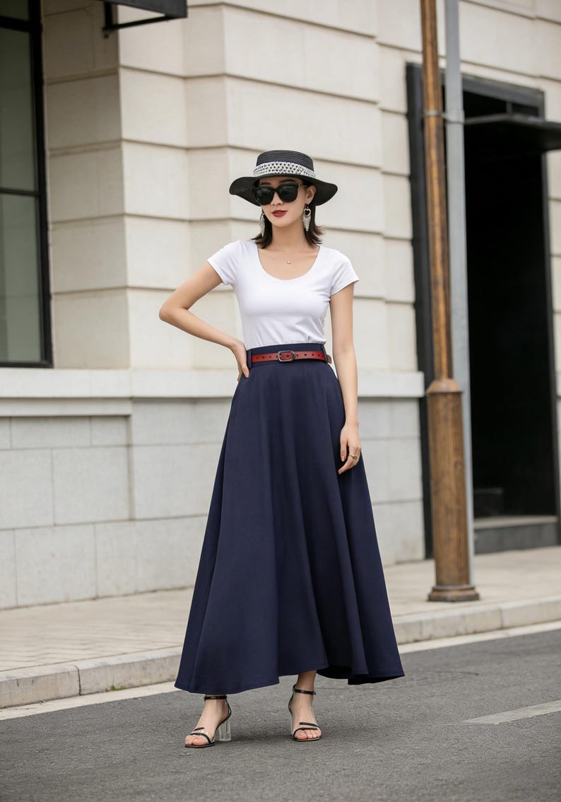 May include: A woman's outfit featuring a white scoop-neck t-shirt, a navy blue maxi skirt, and a brown belt. The outfit is accessorized with a black hat, sunglasses, and sandals. The skirt has a flowing, A-line silhouette.