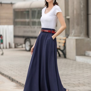 May include: A woman wearing a white short-sleeve top and a long navy blue skirt. A red belt cinches the waist. She is wearing a black hat, sunglasses, and sandals. The outfit is suitable for warm weather.