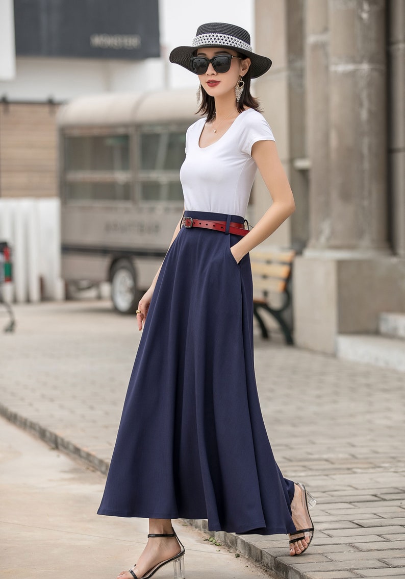 May include: A woman wearing a white short-sleeve top and a long navy blue skirt. A red belt cinches the waist. She is wearing a black hat, sunglasses, and sandals. The outfit is suitable for warm weather.