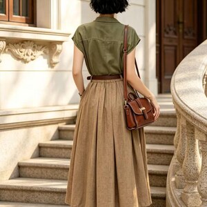 May include: A woman in a stylish outfit stands on outdoor stairs. She wears a short-sleeved olive green button-up shirt, a brown pleated skirt, and a brown leather belt. A brown leather purse hangs from her shoulder. Black high heels complete the look.