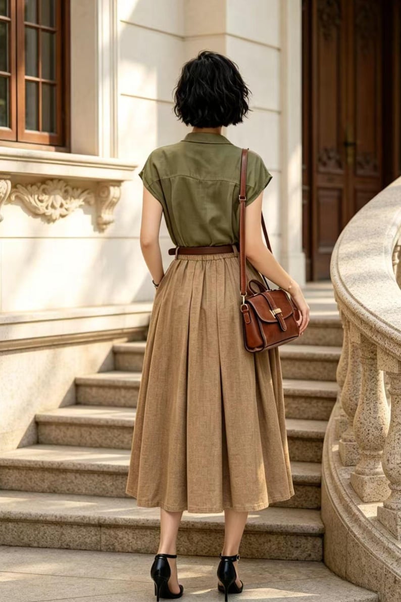 May include: A woman in a stylish outfit stands on outdoor stairs. She wears a short-sleeved olive green button-up shirt, a brown pleated skirt, and a brown leather belt. A brown leather purse hangs from her shoulder. Black high heels complete the look.