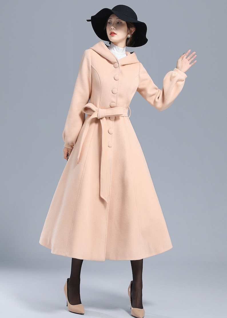 May include: A long, light pink coat with a hood, a belt, and a row of buttons down the front. The coat has a flared skirt and long sleeves.