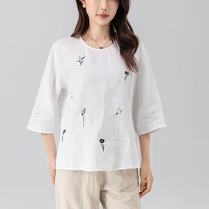 May include: White linen top with embroidered floral details. The top has a round neckline and three-quarter sleeves. The model is wearing beige pants. This casual blouse is suitable for everyday wear.