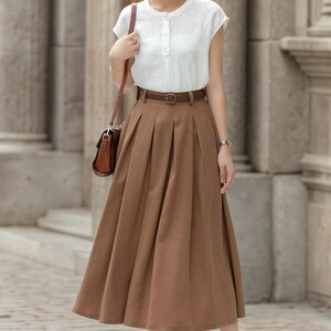 May include: A woman wearing a white short-sleeved blouse and a brown pleated midi skirt. The outfit is accessorized with a brown belt, a brown shoulder bag, and black sandals. The background features stone columns.