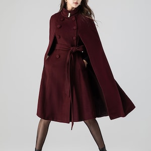 Pu&ograve; includere: Un cappotto a mantella color bordeaux scuro con collo alto a volant e un dettaglio a perla, chiusura doppiopetto e cintura in tessuto abbinata. Il capo presenta aperture per le braccia e una silhouette svasata sotto le ginocchia.