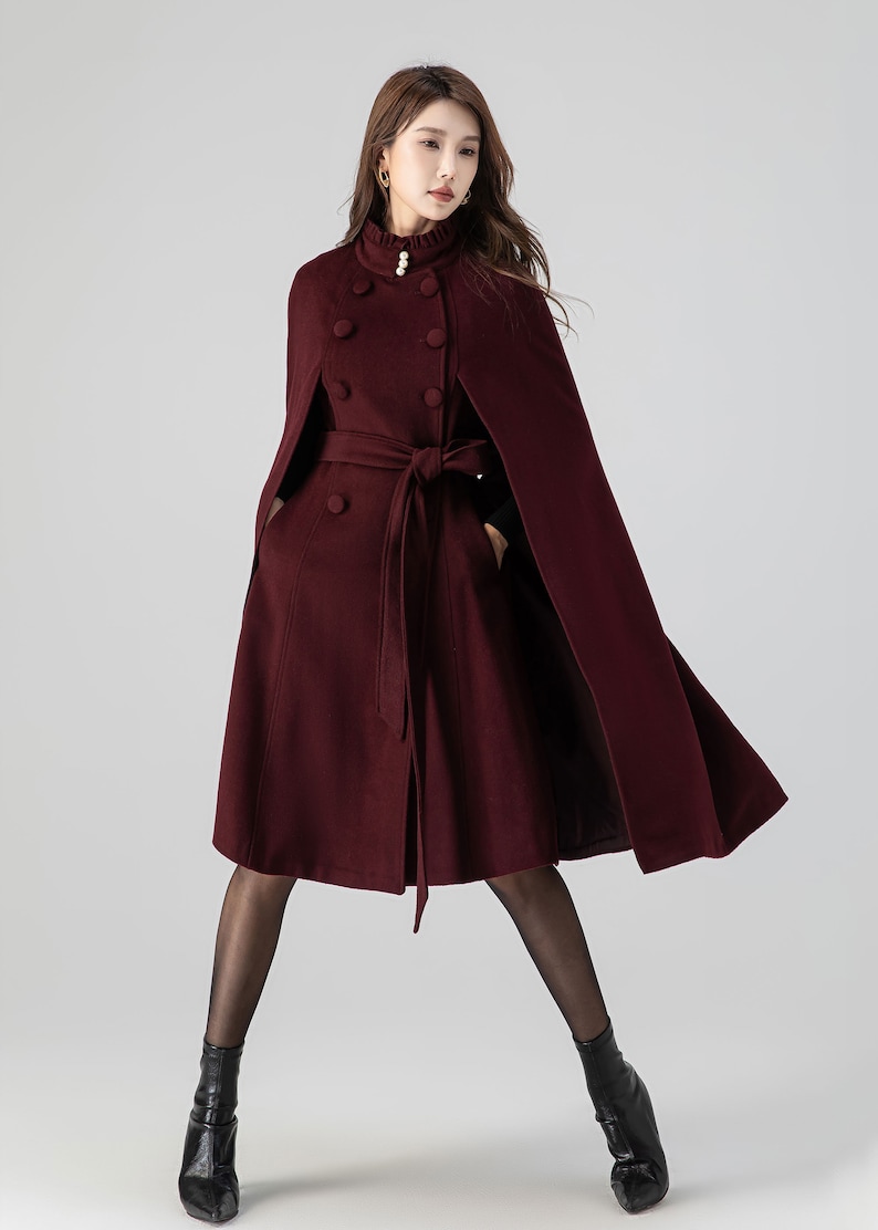 Pu&ograve; includere: Un cappotto a mantella color bordeaux scuro con collo alto a volant e un dettaglio a perla, chiusura doppiopetto e cintura in tessuto abbinata. Il capo presenta aperture per le braccia e una silhouette svasata sotto le ginocchia.