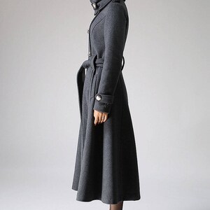 May include: A long, gray wool coat with a high collar, a belt, and button details. The coat has a flared skirt and is worn with black heeled boots.