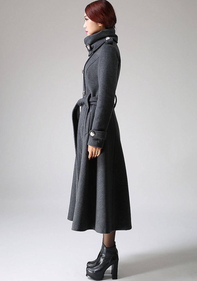 May include: A long, gray wool coat with a high collar, a belt, and button details. The coat has a flared skirt and is worn with black heeled boots.
