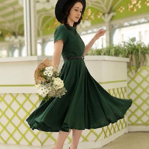 Puede incluir: Un vestido midi de color verde oscuro con mangas cortas y falda amplia, combinado con un cintur&oacute;n negro fino, un sombrero de paja negro y zapatos de tac&oacute;n beige con tira en T. Se sostiene un ramo de flores blancas envuelto en papel.